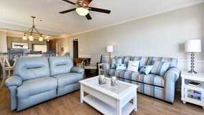 Un salon avec un canapé bleu, un canapé à carreaux, une table basse blanche et un ventilateur de plafond.