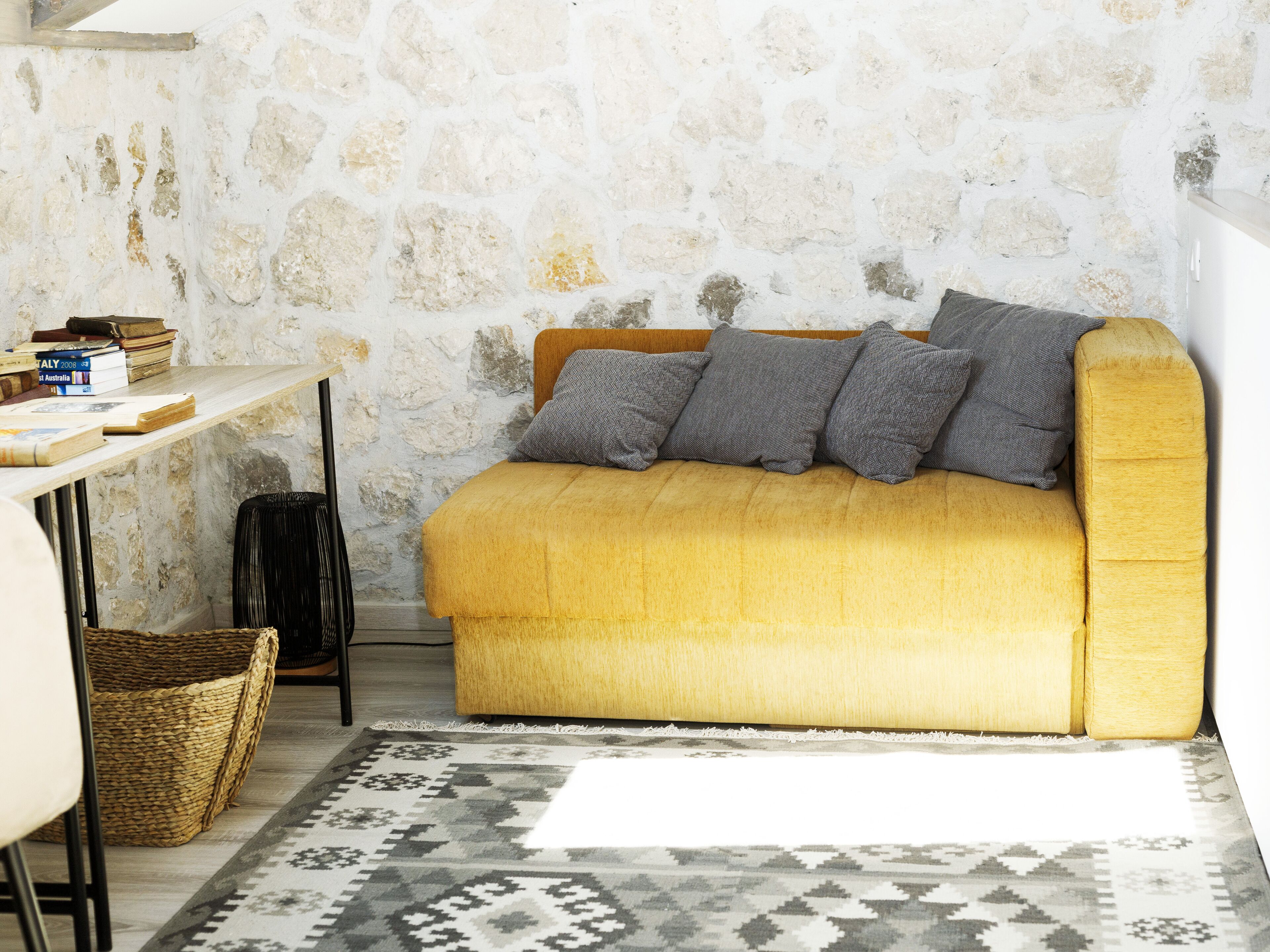 A cozy room with a yellow sofa, a stone wall, a small table with books, and a patterned rug.