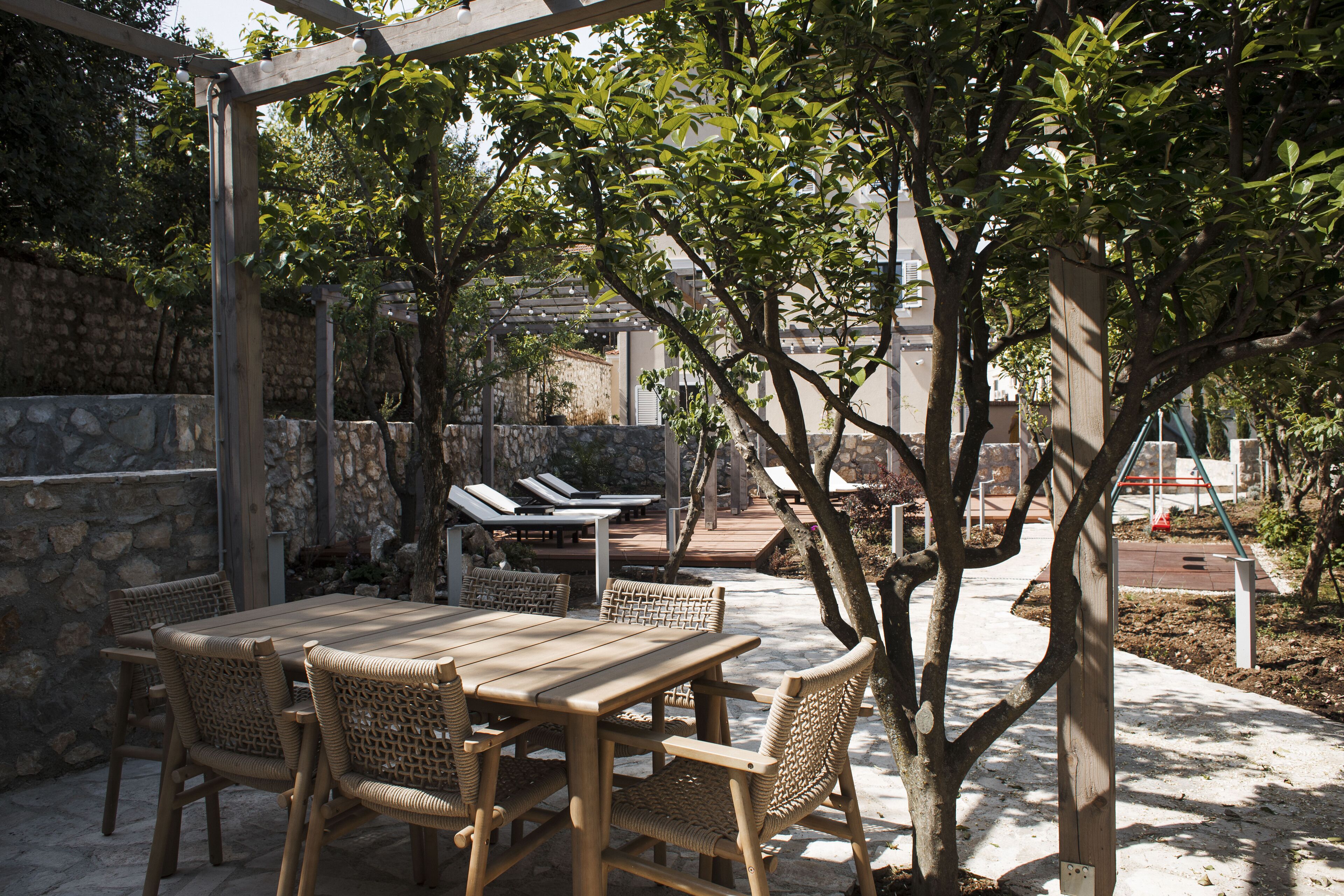 An outdoor patio with a wooden table and wicker chairs, surrounded by stone walls and trees.