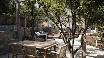 An outdoor patio with a wooden table and wicker chairs, surrounded by stone walls and trees.
