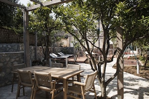 Un patio al aire libre con mesa de madera y sillas de mimbre, rodeado de muros de piedra y árboles.