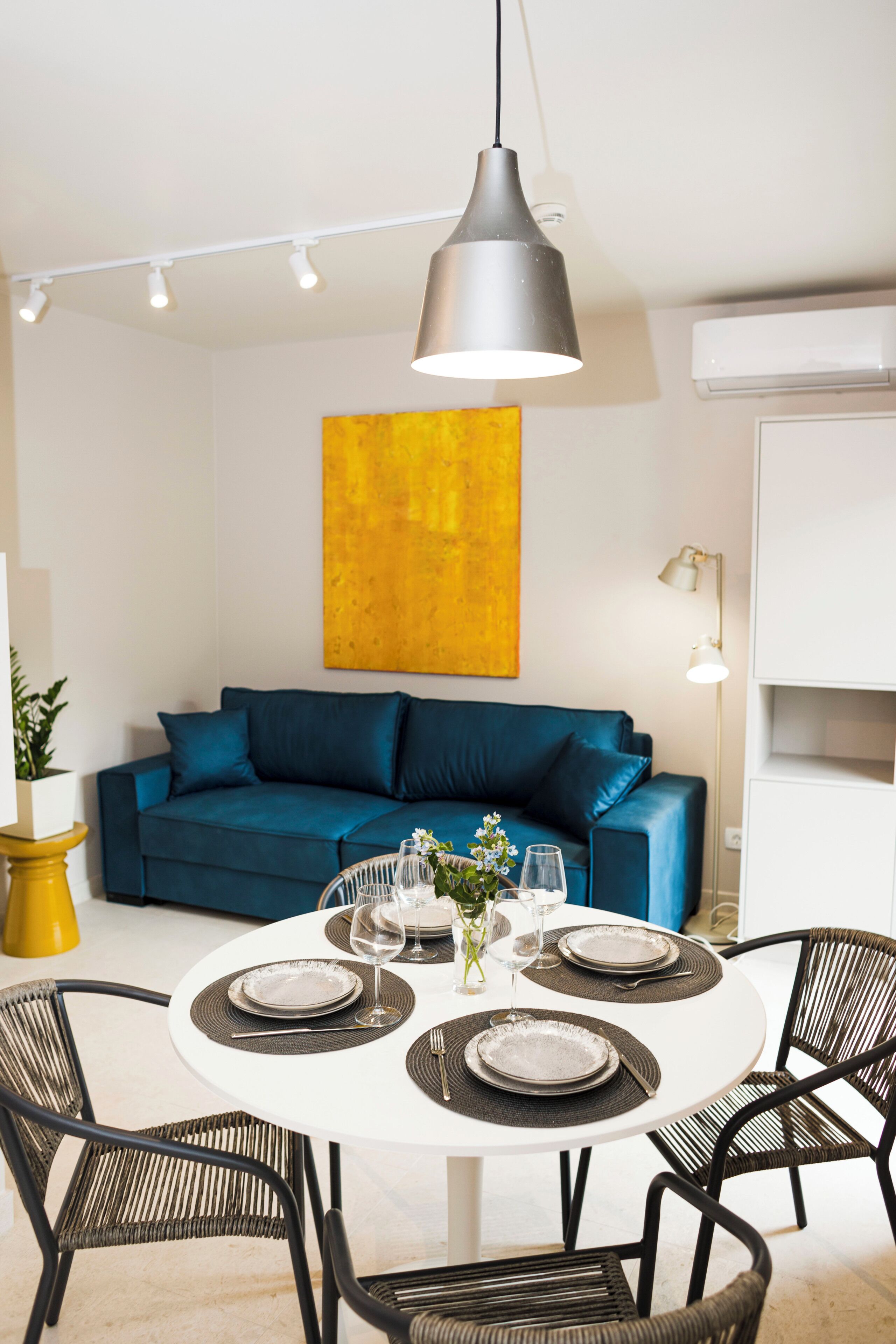 A dining area with a round table set for four, a blue sofa, a yellow accent chair, and a modern pendant light.