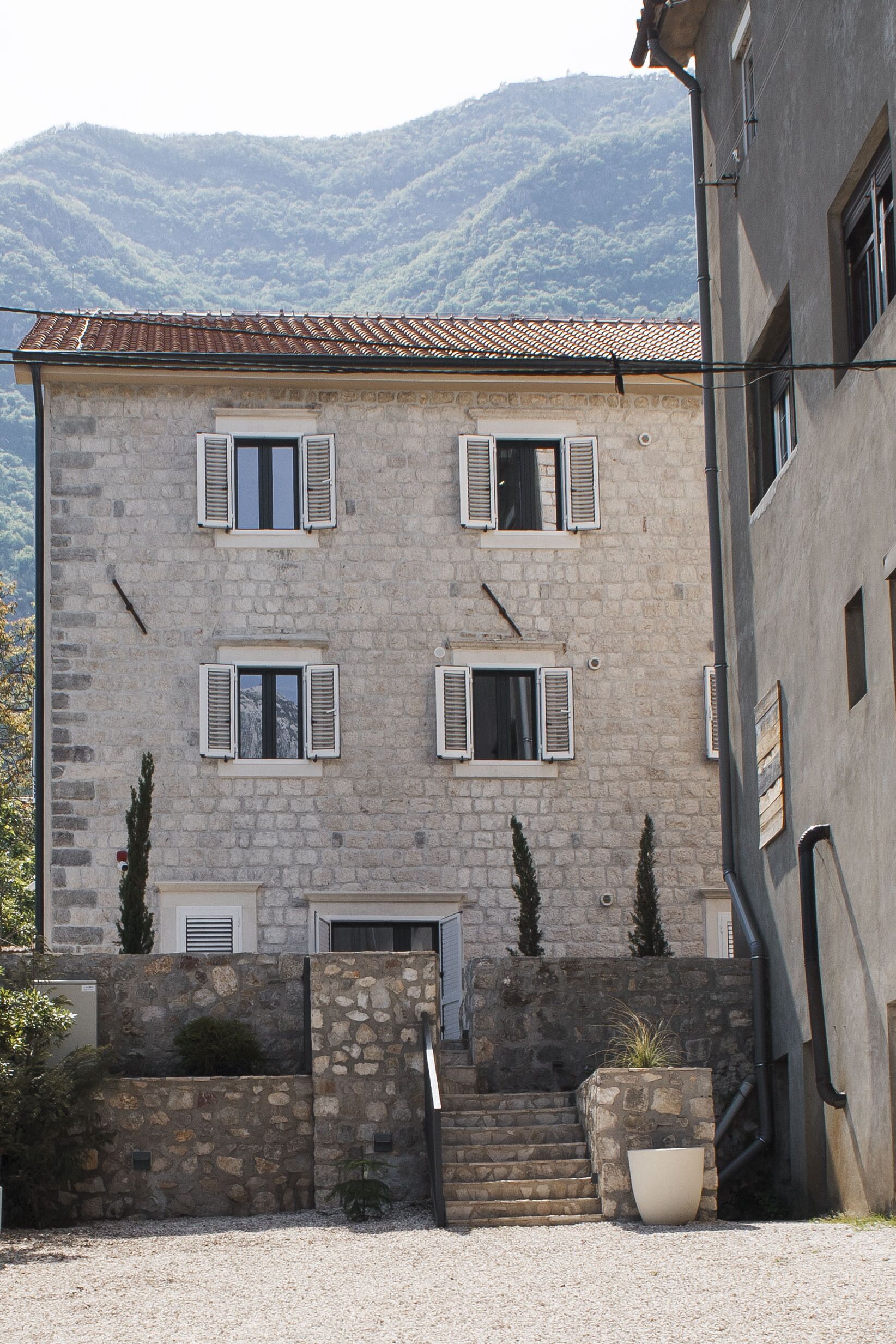 A stone building with a tiled roof, multiple windows with shutters, and a staircase leading to the entrance.