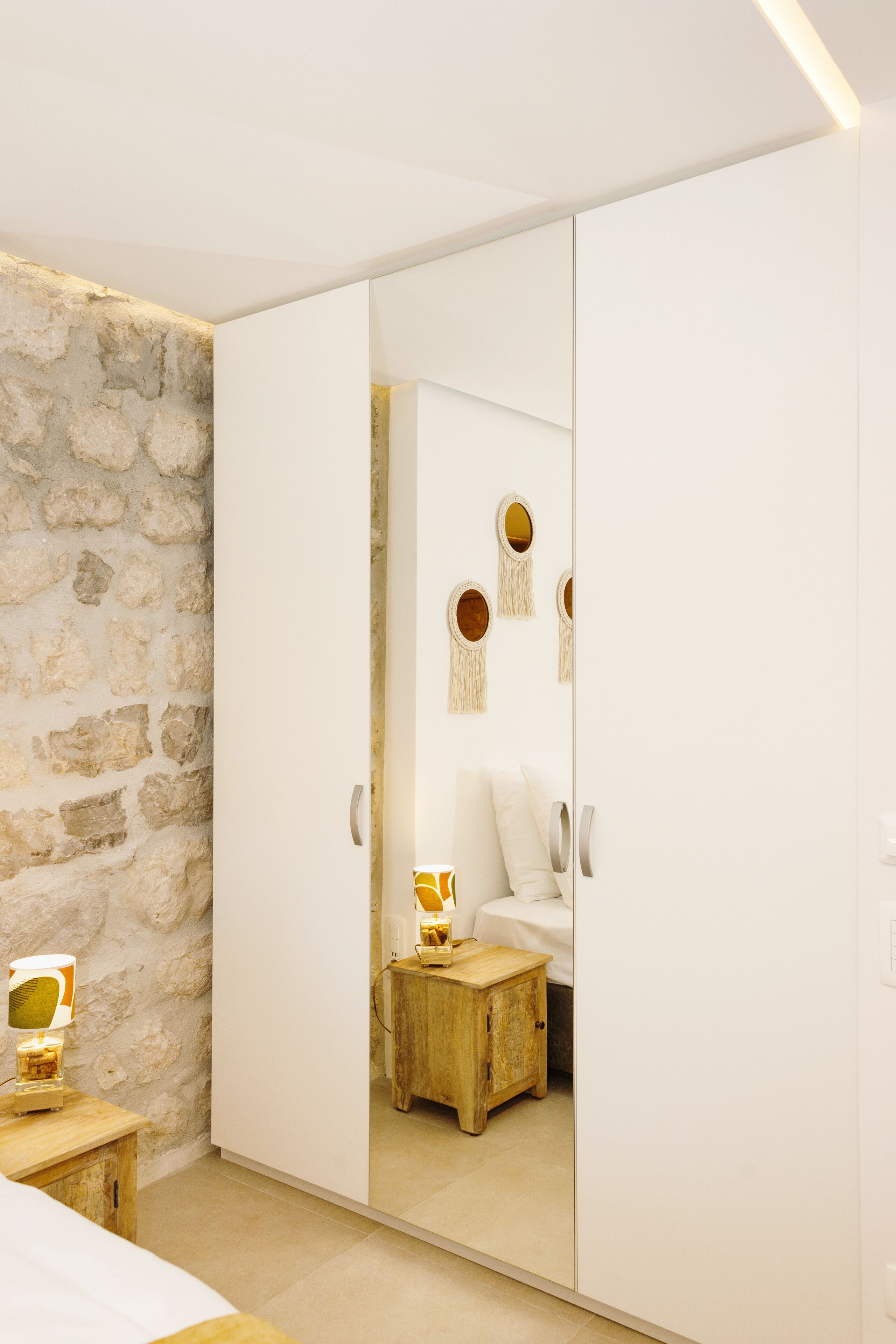 A bedroom with a stone wall, a wooden nightstand, a bed, and a wardrobe with a mirror.