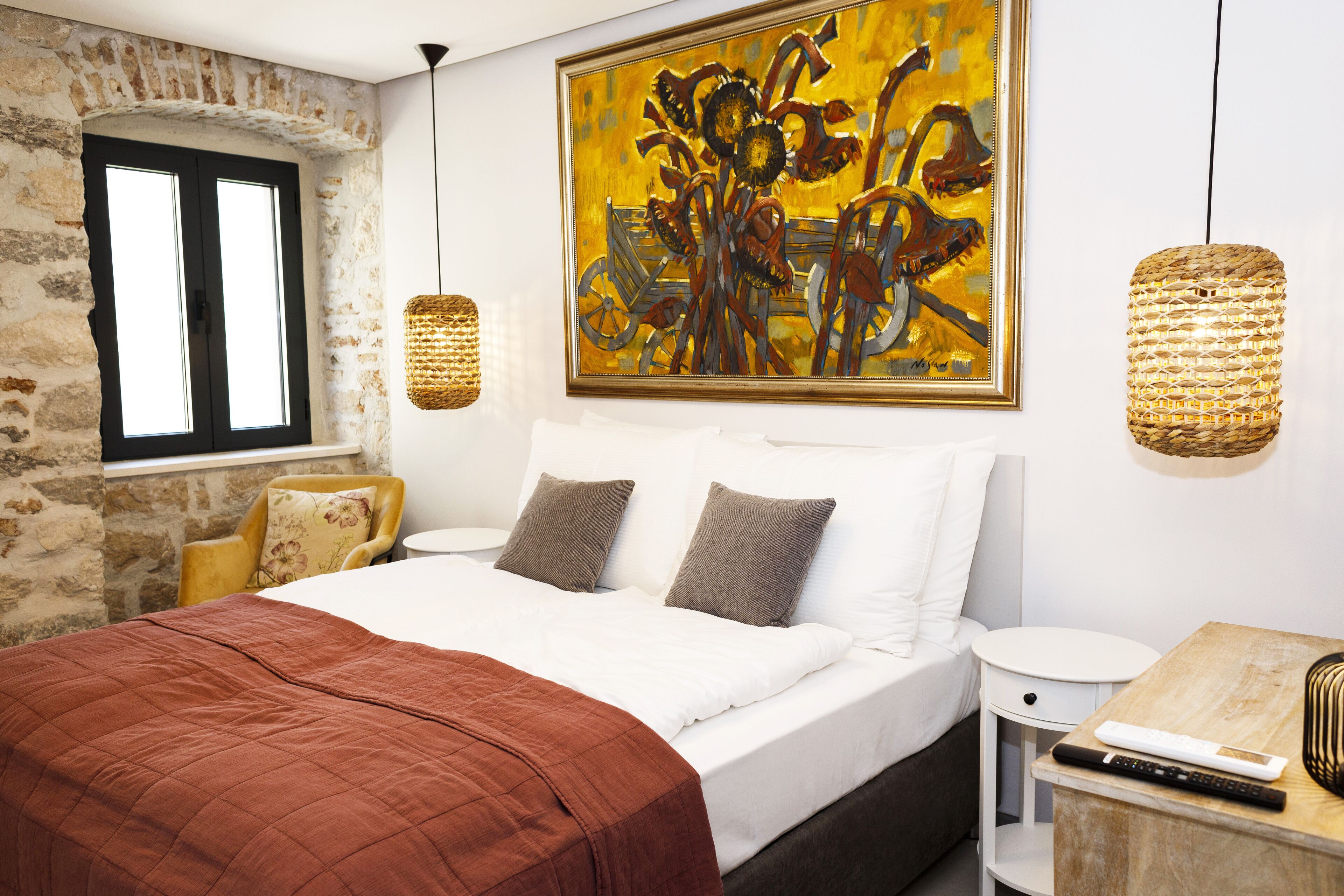 A bedroom with a large bed, a stone wall, a window, a painting, and a woven light fixture.