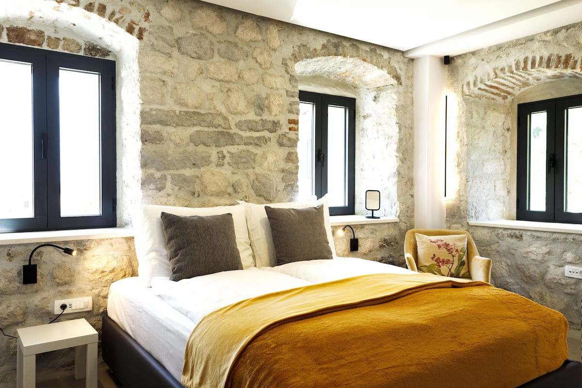 A bedroom with a stone wall, a bed with white bedding, and a window with a black frame.