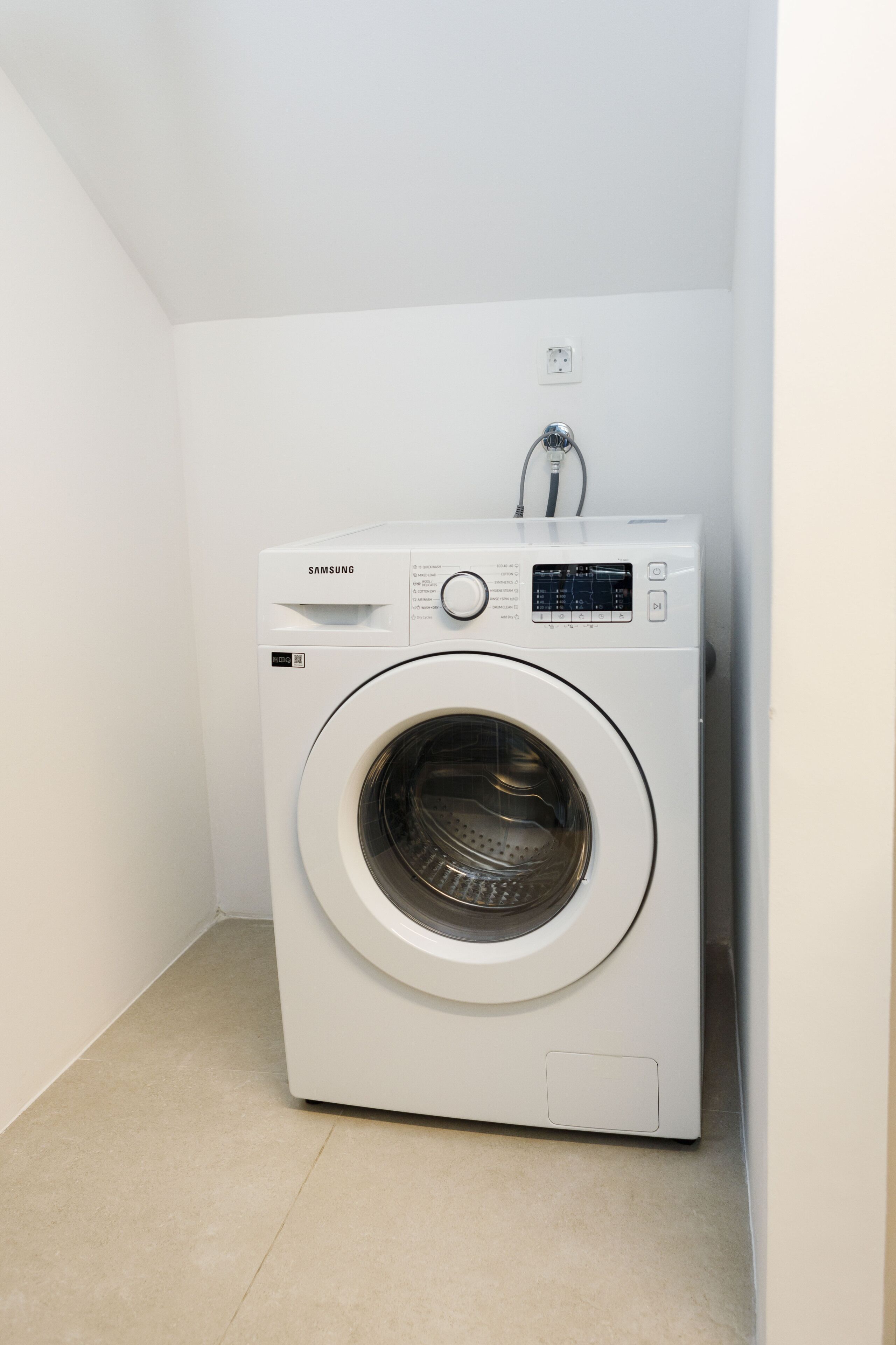A white Samsung washing machine in a corner of a room with white walls and a tiled floor.