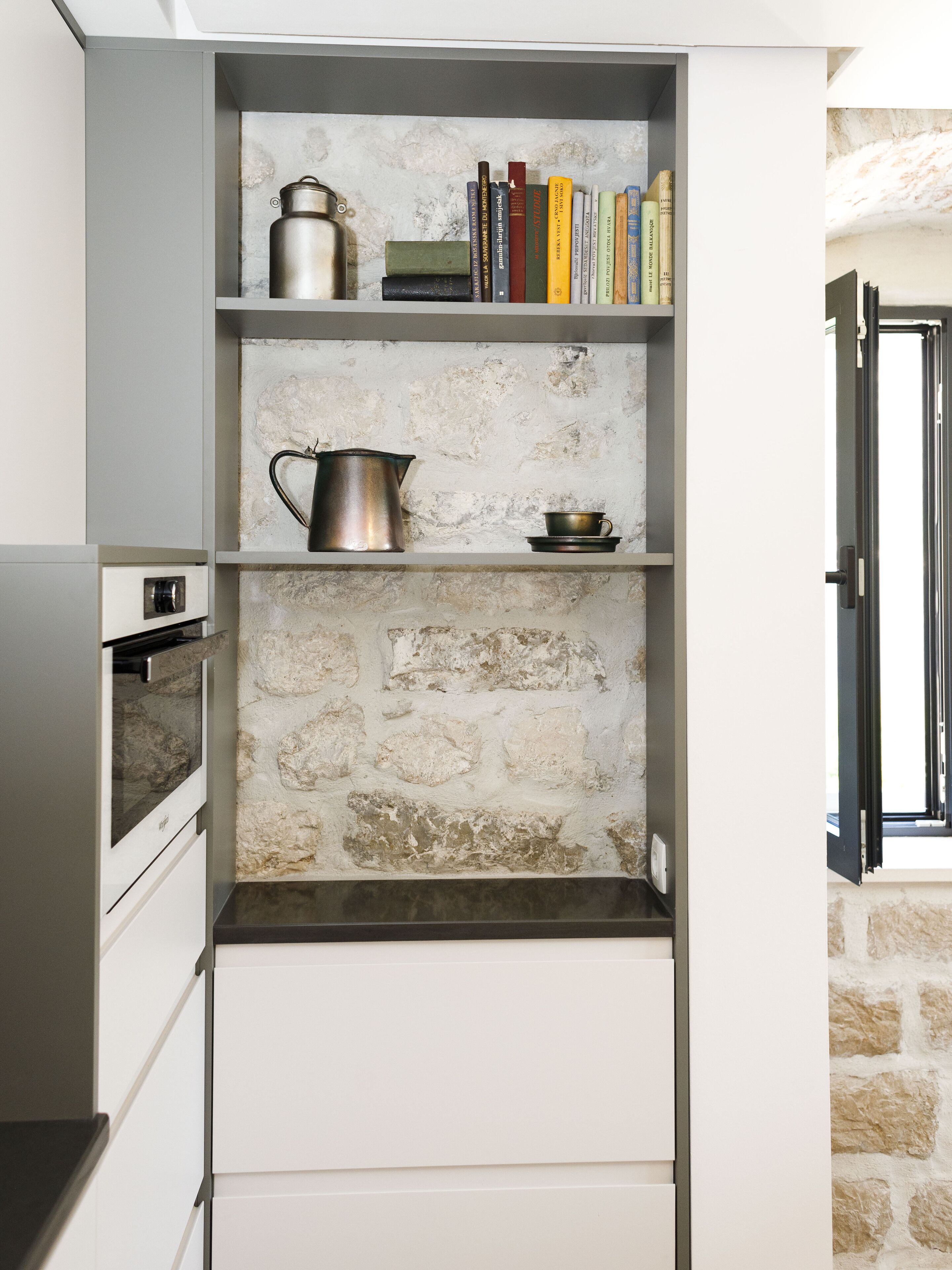 A kitchen with a built-in oven, a coffee maker, a kettle, and a bookshelf with books.