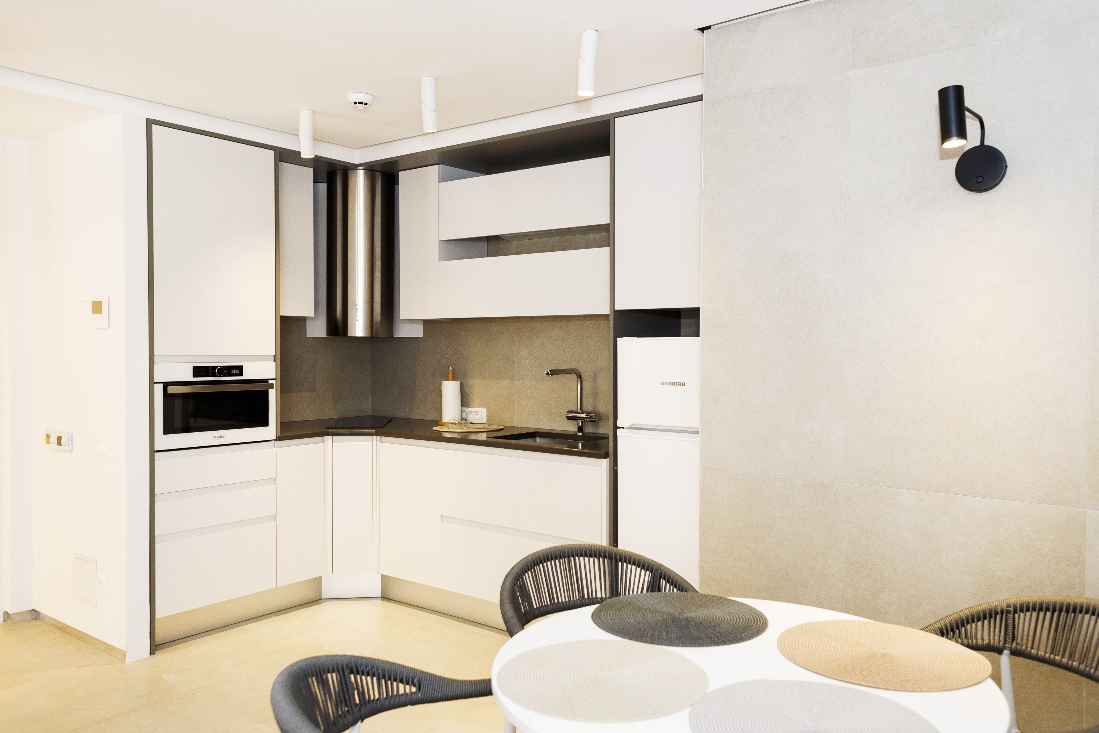 Een moderne keuken met witte kasten, een ingebouwde oven en een zwart aanrecht. Er is een eethoek met een ronde tafel en zwarte stoelen.