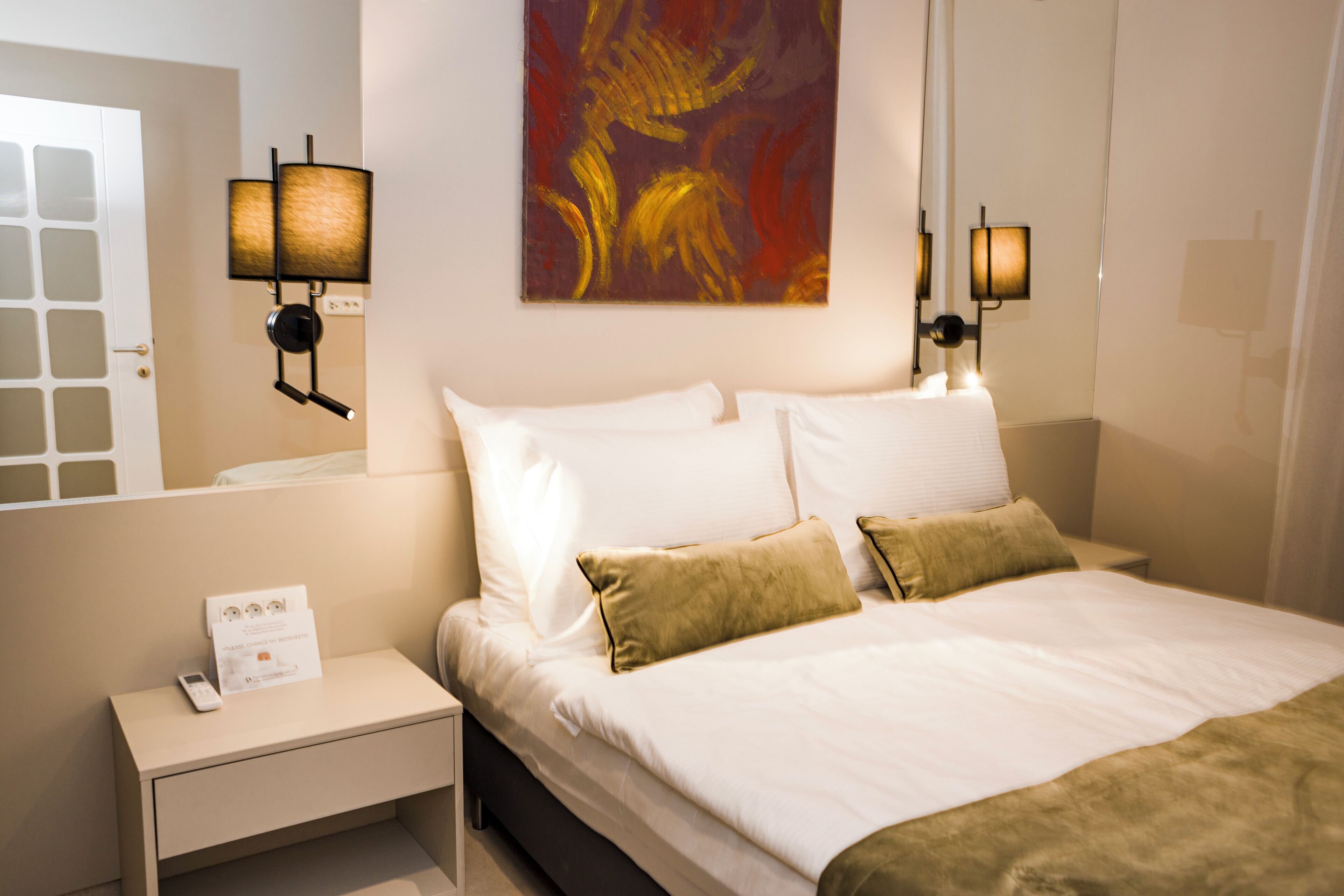 A hotel room with a bed, nightstand, wall-mounted lamps, and a painting.