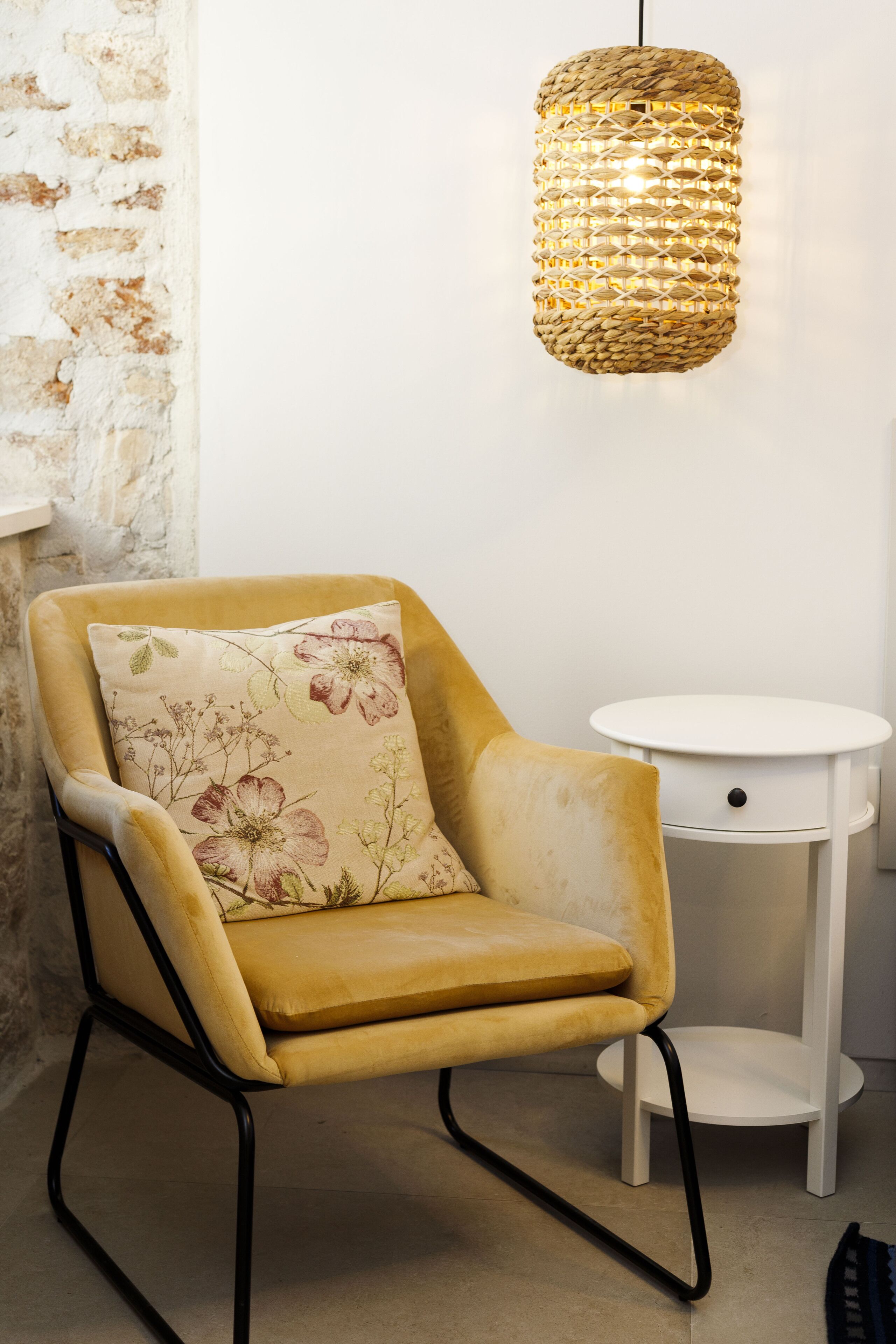 A mustard-colored armchair with a floral pillow, a white round side table, and a woven pendant light.
