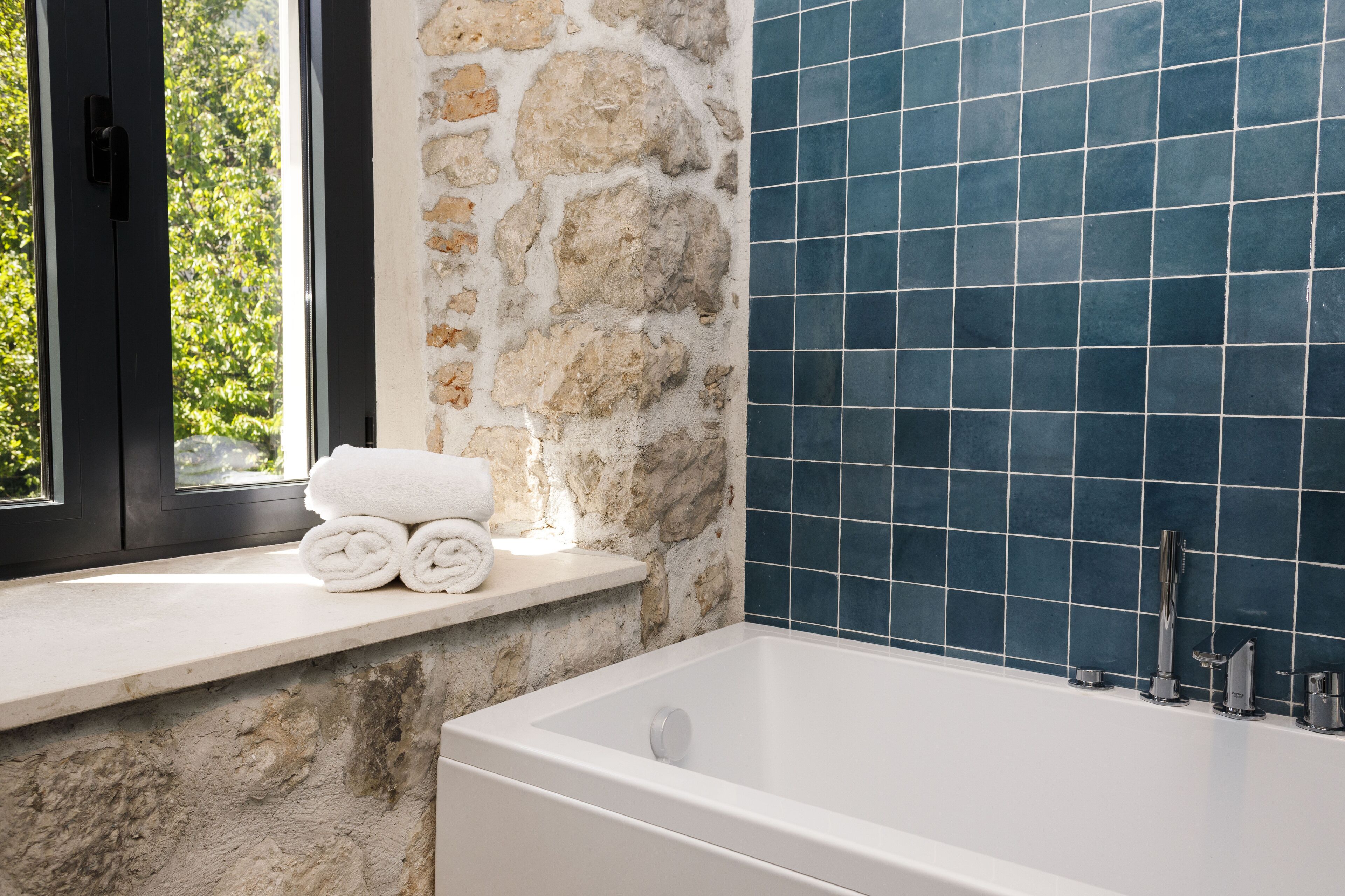 A bathroom with a stone wall, a bathtub, and a window with a view of greenery.