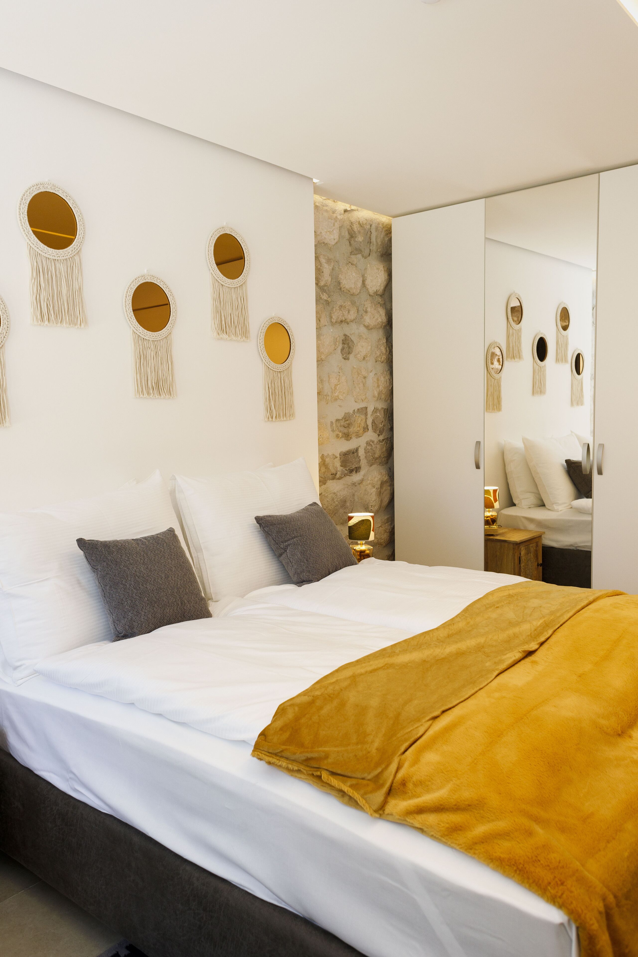 Un dormitorio moderno con cama, ropa de cama blanca, pared decorada con piedra y adornos de macramé colgados en la pared.