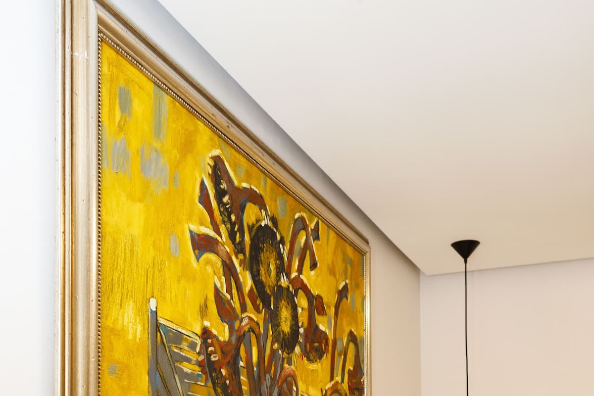 A room with a large painting of a bicycle and flowers, a bed with white and gray pillows, and a hanging woven light fixture.
