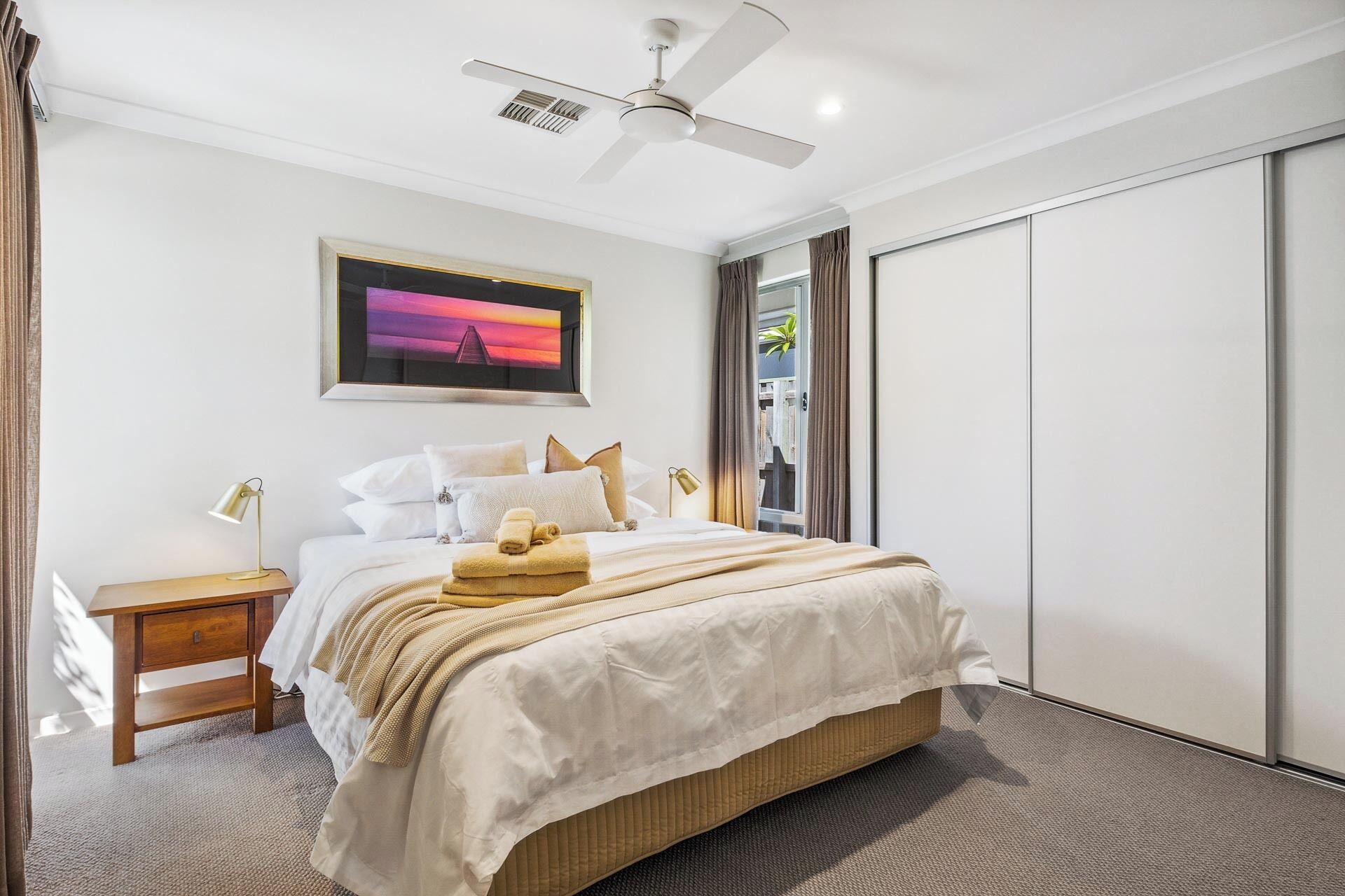 A bedroom with a bed, bedside tables, a ceiling fan, a framed picture, and a sliding door.