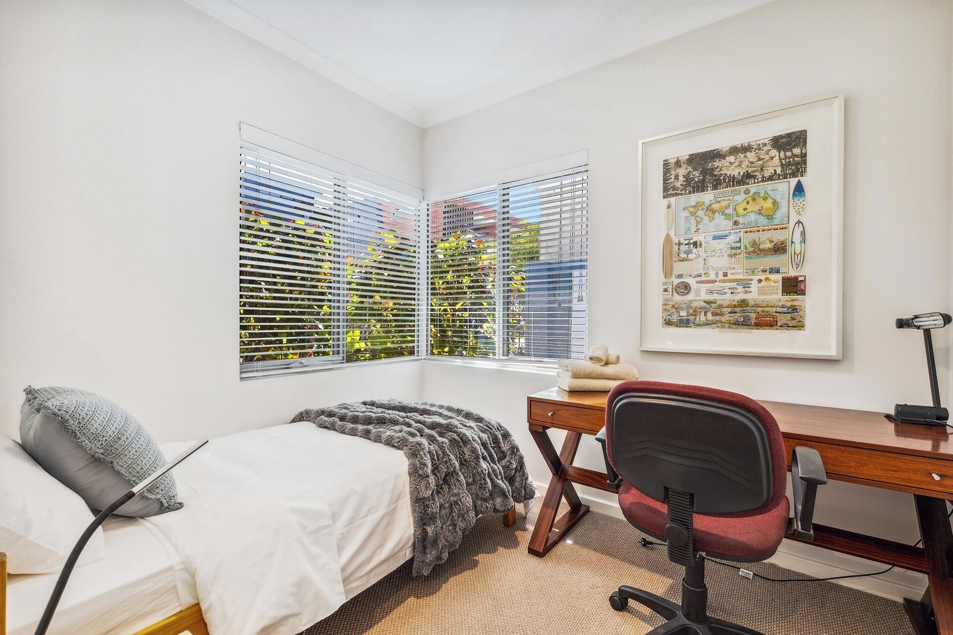 A bedroom with a bed, a desk, and a chair. There are two windows with blinds, and a framed map on the wall.