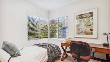 A bedroom with a bed, a desk, and a chair. There are two windows with blinds, and a framed map on the wall.