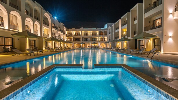 A swimming pool with lounge chairs and umbrellas, surrounded by multi-story buildings with balconies. - Al Masa Hotel El Sokhna (Ataqah)