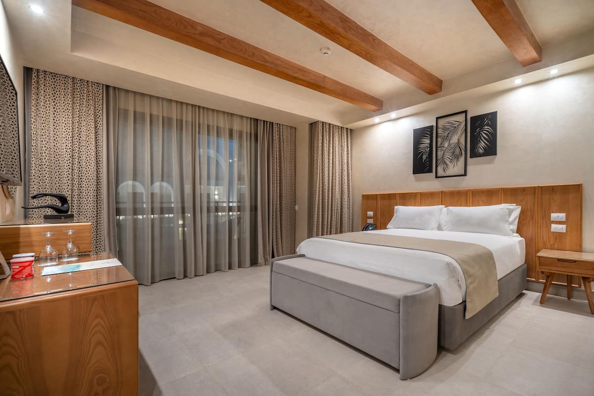 Une chambre d’hôtel moderne avec un grand lit, une tête de lit en bois, une table de chevet, un banc et un bureau équipé d’un ordinateur et d’un téléphone.