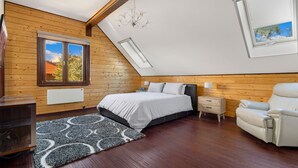 A bedroom with a sloped ceiling, wooden walls, a bed, a nightstand, a chair, and a window with a view of trees. - Deepwater - 68 Kunama Drive (East Jindabyne)
