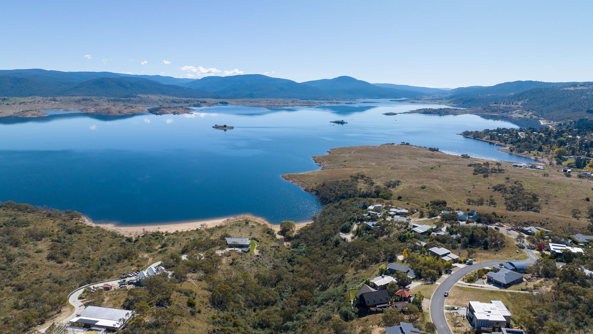 An aerial view of a lakeside community with residential buildings, roads, and a large body of water surrounded by hills.