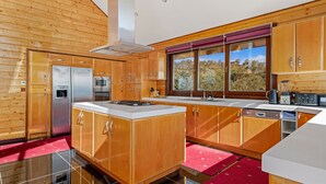 A modern kitchen with wooden cabinetry, stainless steel appliances, and a large window offering a view of trees. - Deepwater - 68 Kunama Drive (East Jindabyne)