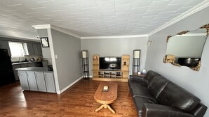 A modern living room with a black leather sofa, wooden coffee table, and a flat-screen TV. A kitchen with grey cabinets and a black refrigerator is visible in the background. - Quiet Country style living, your home away from home.  (Stephenville)
