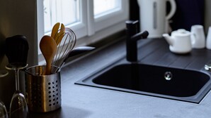 Una cocina con un lavamanos de acero inoxidable, un hervidor y una ventana con vista a flores.