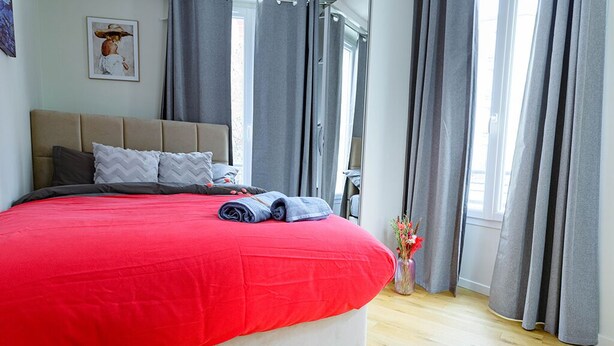 Un dormitorio con una cama grande, cortinas grises, una ventana y un jarrón con flores.