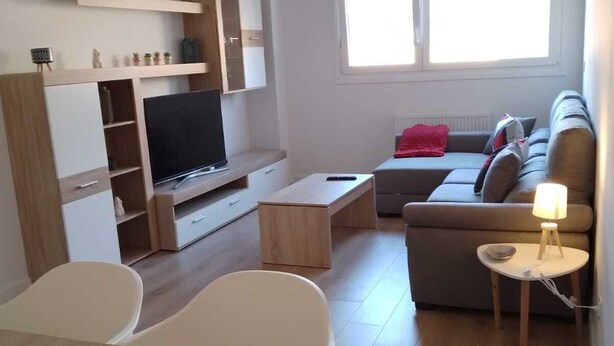 A modern living room with a flat-screen TV, a sofa, a dining table, and a small side table with a lamp. - Apartamento Pamplona (Pamplona)