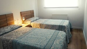 A bedroom with two beds, a bedside table with a lamp, and a window with blinds. - Apartamento Pamplona (Pamplona)