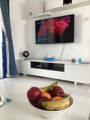 Un moderno salón con un televisor de pantalla plana montado en la pared, un mueble blanco de entretenimiento debajo y un frutero sobre la mesa.
