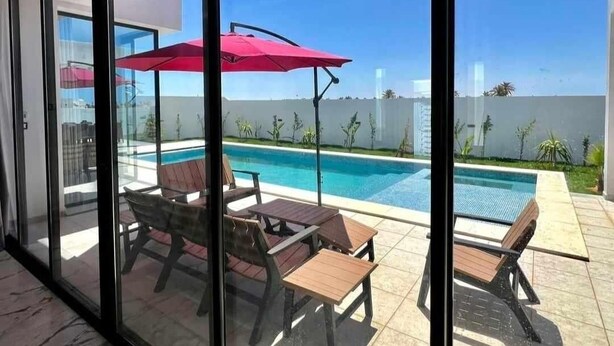 Un área de piscina exterior moderna con puertas de cristal, sillas de lounge de madera y un paraguas rojo.