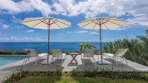 Eine Terrasse mit zwei weißen Liegestühlen und zwei großen Sonnenschirmen, mit Blick auf einen Pool und das Meer.