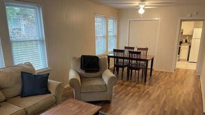 Ein Wohnzimmer mit Sofa, Sessel, Couchtisch, Fernseher, Esstisch, Stühlen und einer Küche im Hintergrund.