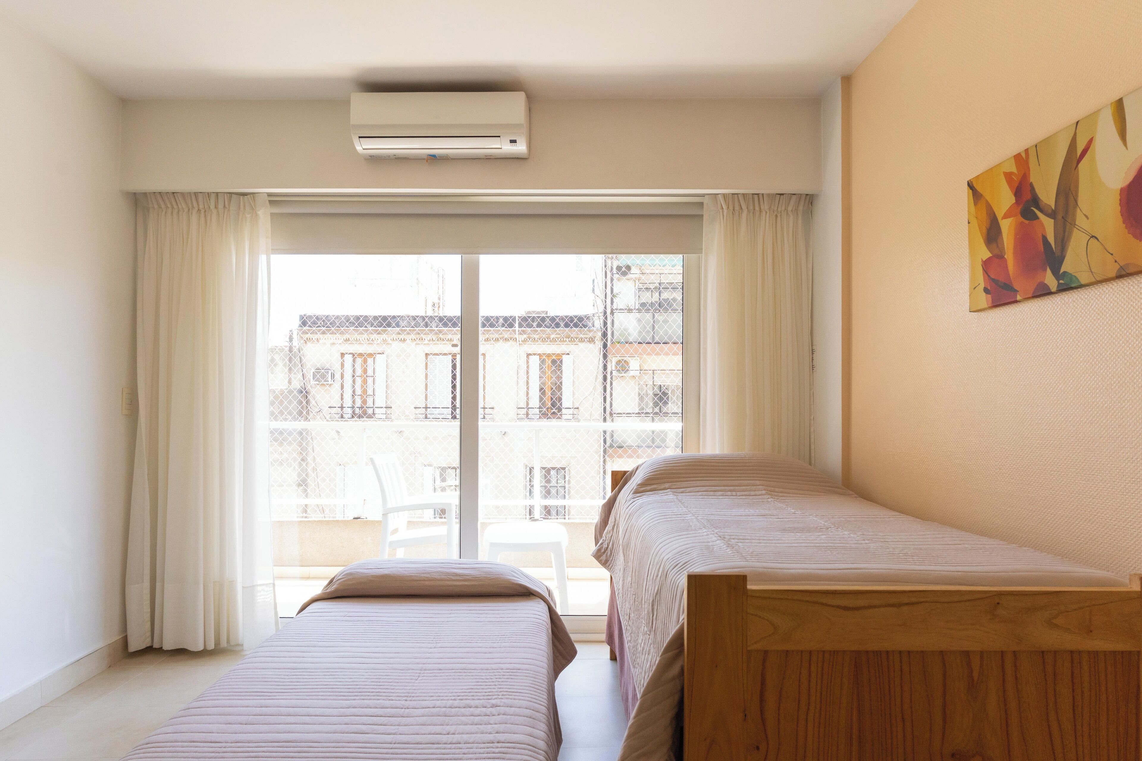Een slaapkamer met twee bedden, een raam met gordijnen en uitzicht op gebouwen.