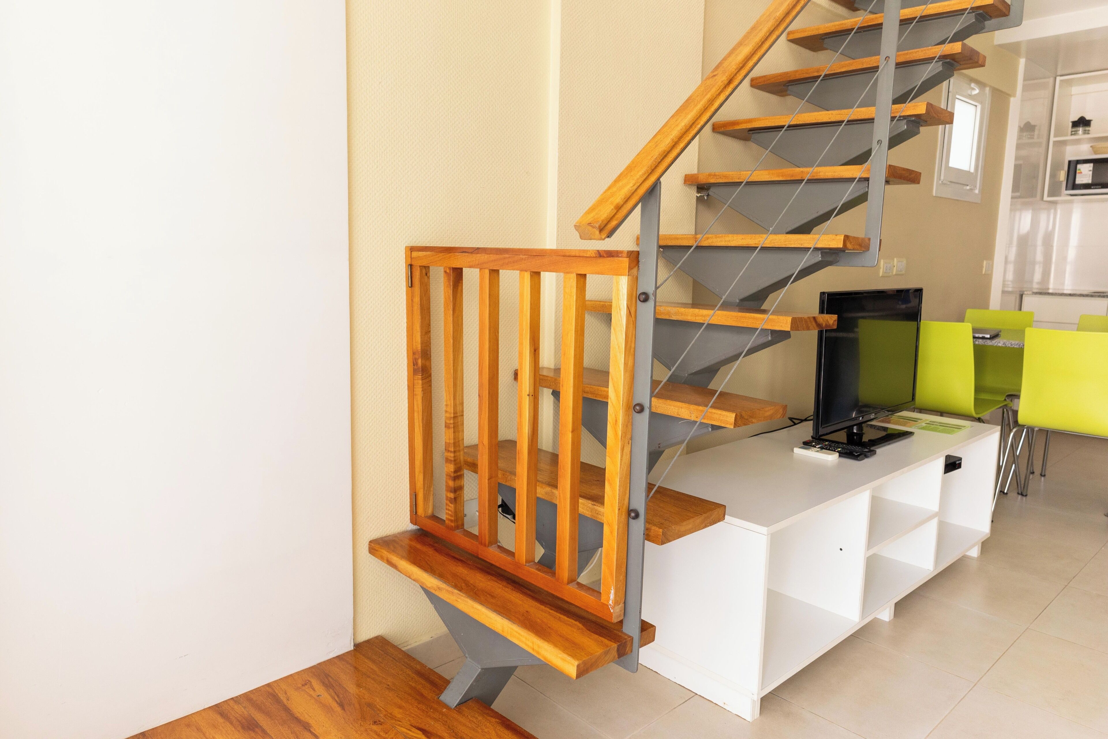 Una moderna scala con gradini in legno e ringhiere metalliche, che conduce a un tavolo consolle bianco con televisore a schermo piatto e sedie verdi.