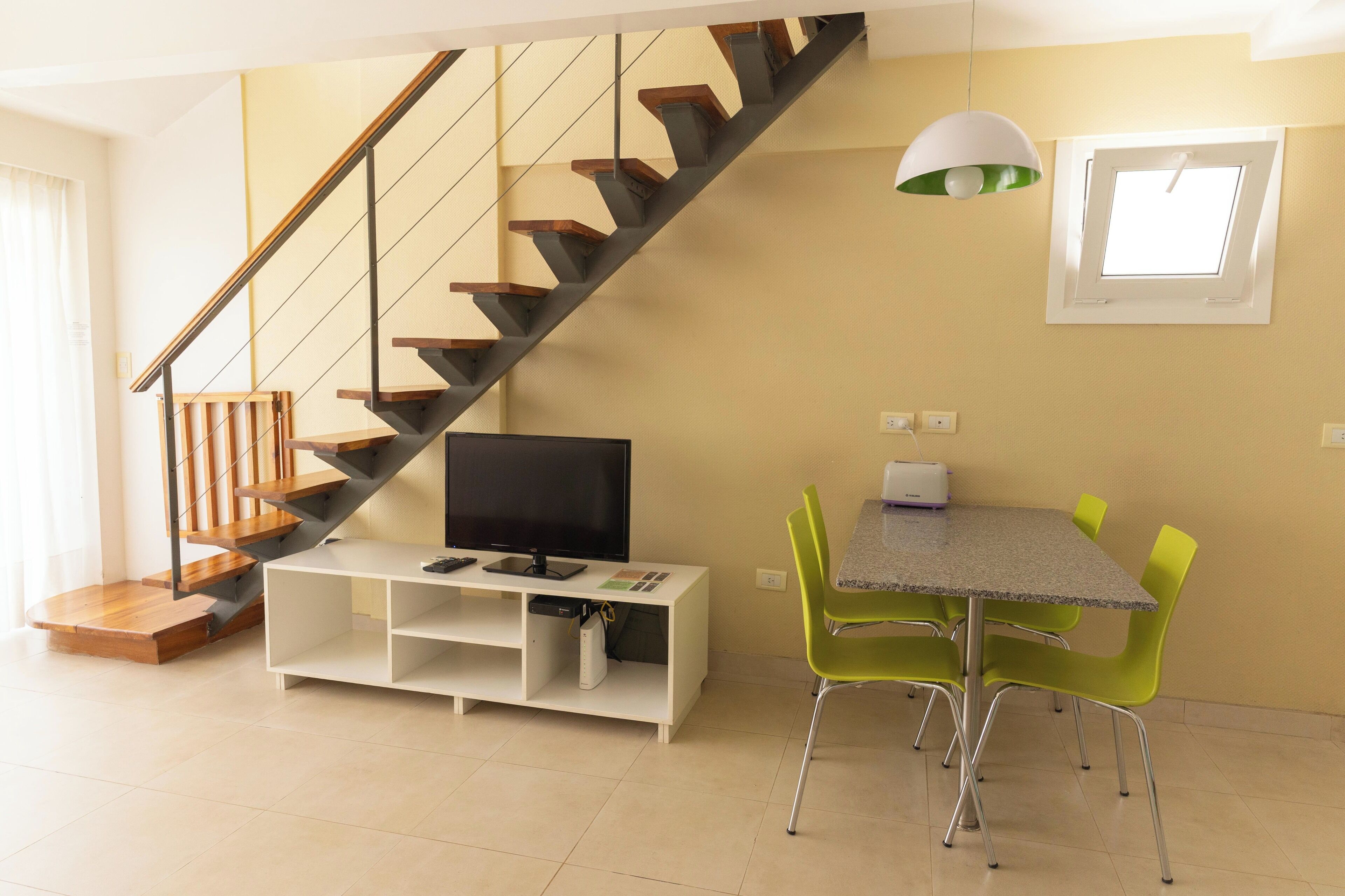 A modern living room with a wooden staircase, a flat-screen TV, a white entertainment unit, and a dining area with green chairs.