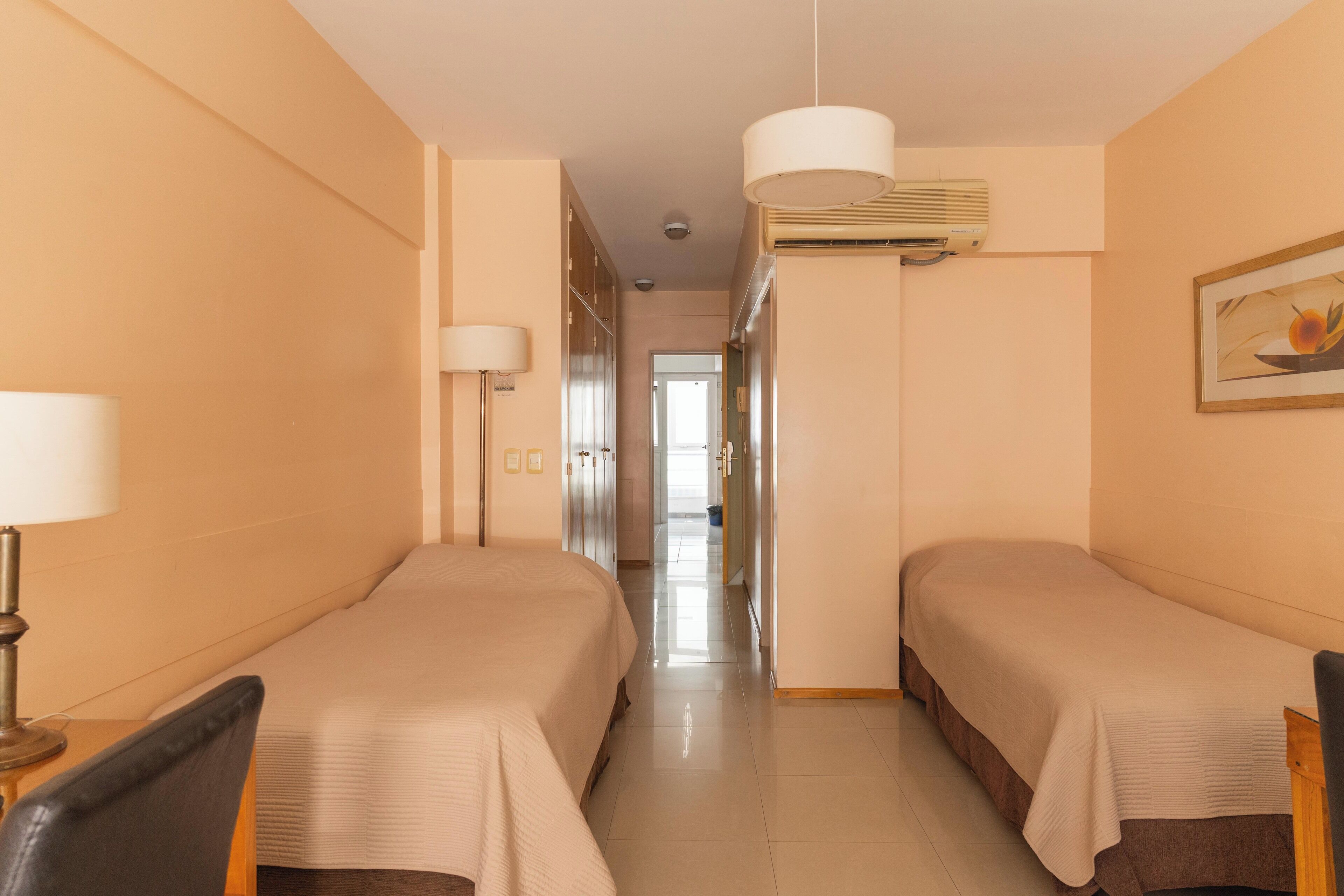 Ett hotellrum med två separata sängar, ett skrivbord, en stol, en lampa, en luftkonditioneringsanläggning och en hall som leder till ett annat rum.