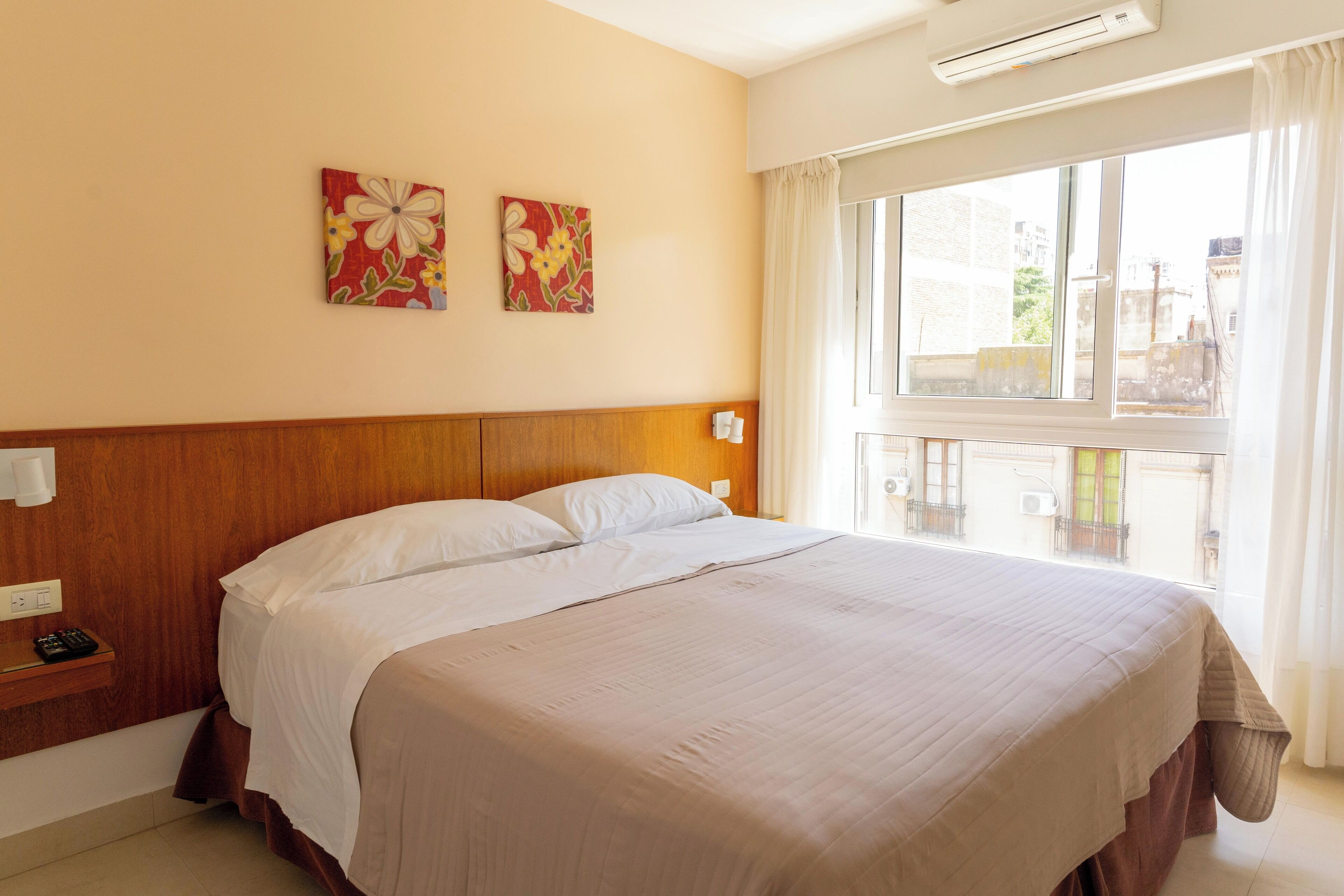 Habitación de hotel con una cama grande, cabecera de madera, dos cuadros florales en la pared, una mesita de noche con un teléfono y una ventana con vistas a edificios.