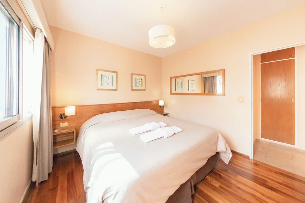 Una habitación de hotel con una cama grande, una mesita de noche con lámpara, un espejo y un armario de madera.