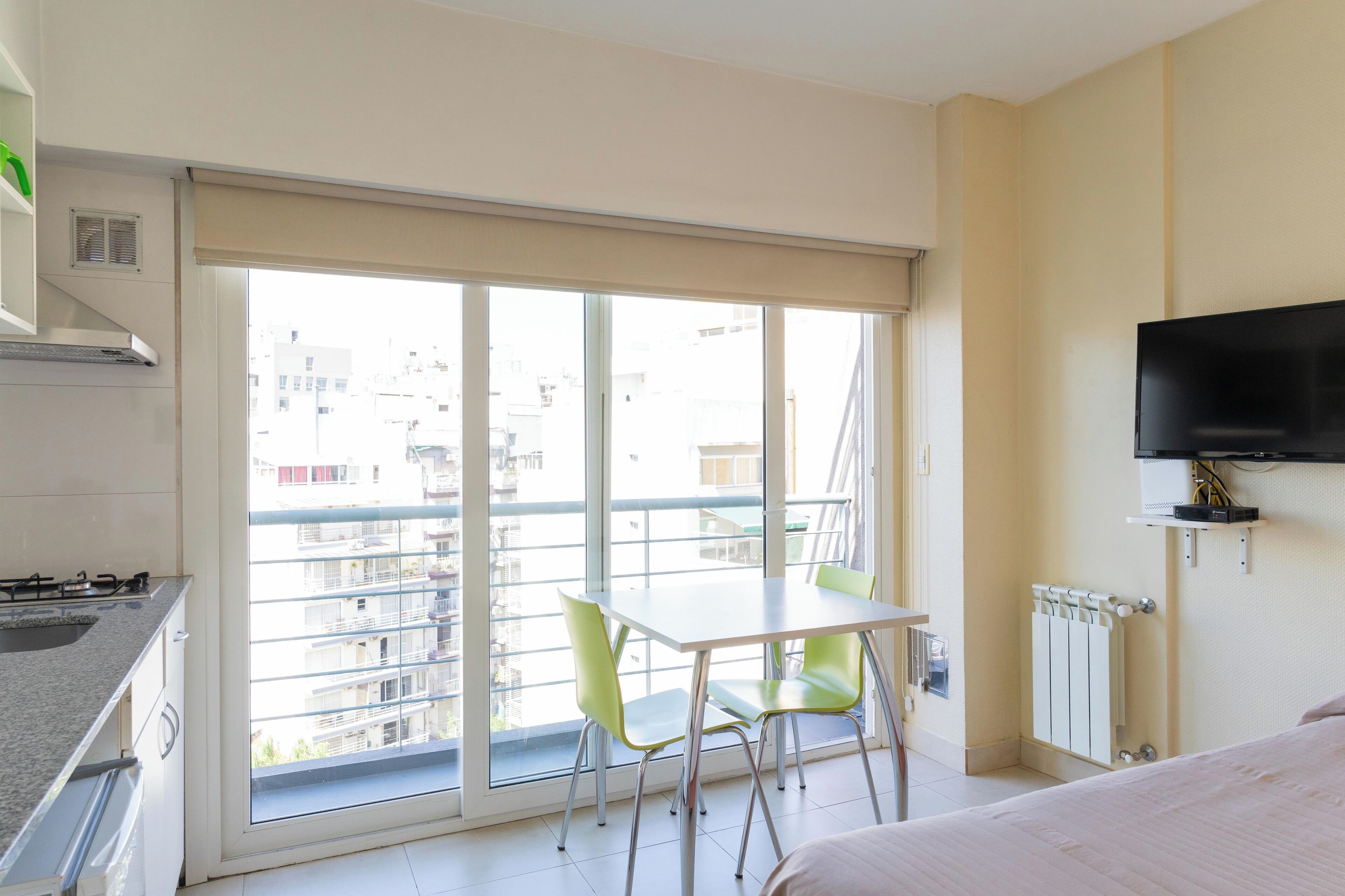 Moderni hotellihuone, jossa on parveke, ruokapöytä tuoleineen, seinälle asennettu televisio ja pieni keittiö.