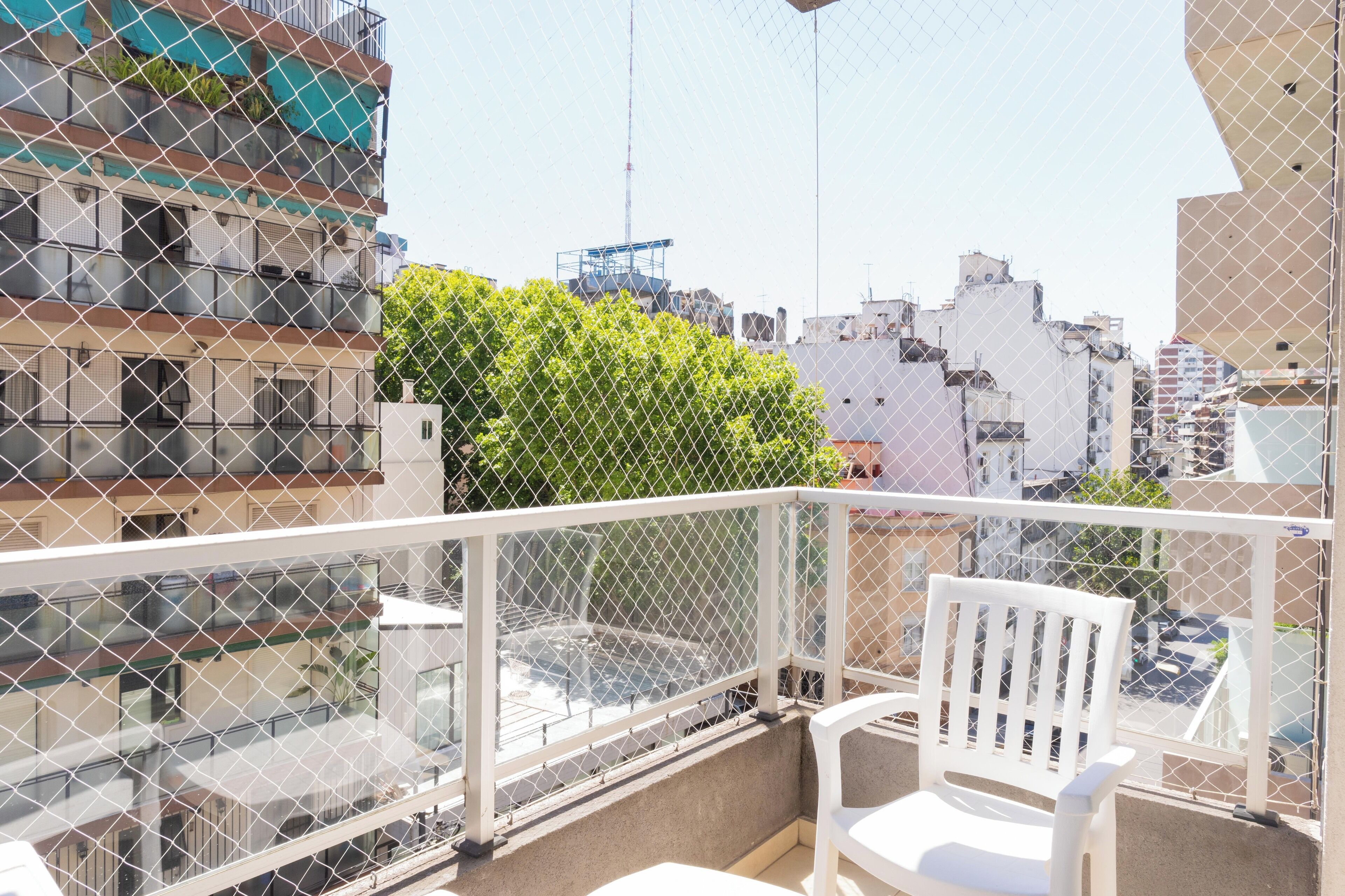 Un balcón con una silla blanca, una cerca de malla y vistas de un paisaje urbano con edificios y vegetación.