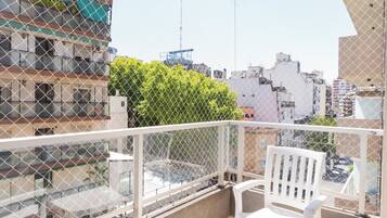 Un balcón con una silla blanca, una cerca de malla y vistas de un paisaje urbano con edificios y vegetación.
