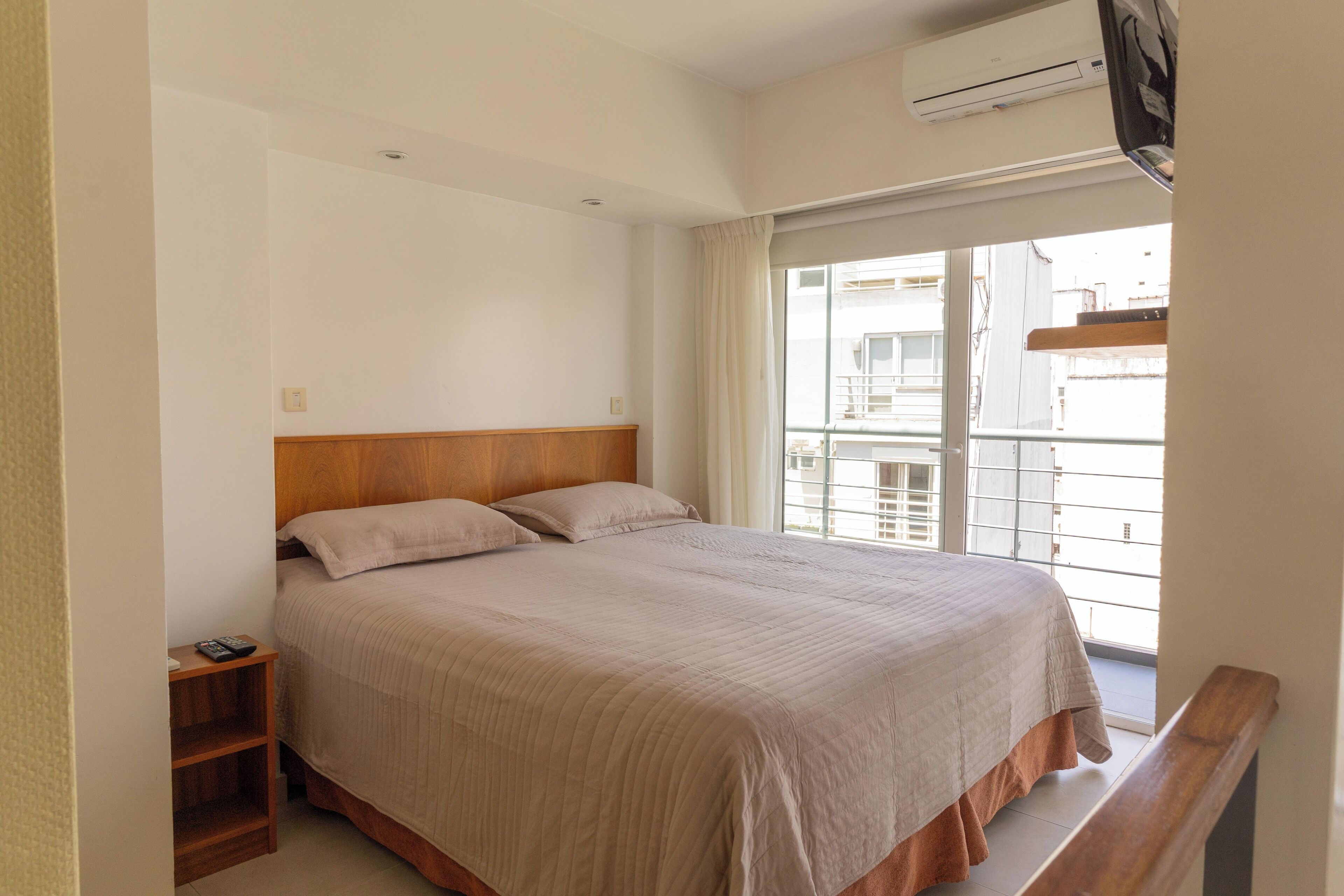 Un dormitorio con cabecera de madera, cama con colcha color beige, mesita de noche con control remoto y ventana con vista a edificios.
