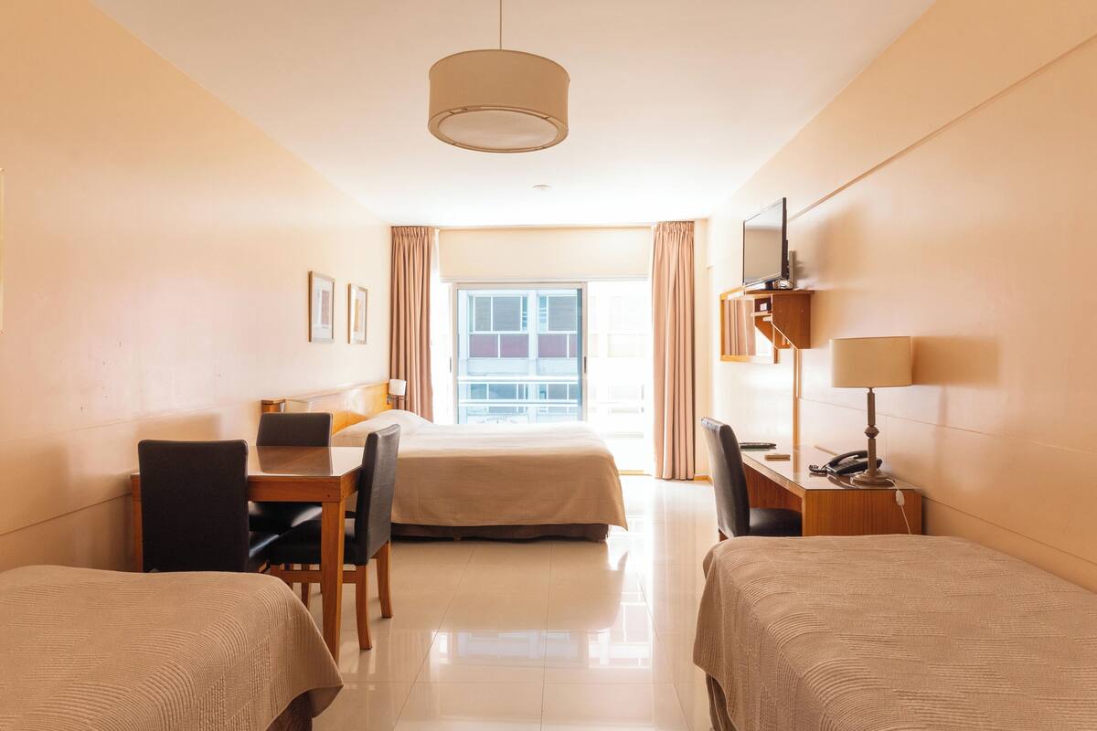Habitación de hotel con dos camas individuales, un escritorio con silla, televisión y ventana con cortinas.