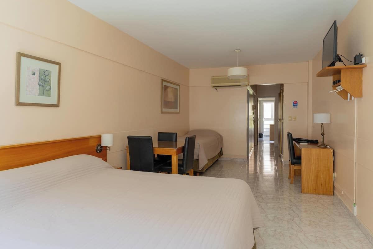Habitación de hotel con una cama grande, un escritorio con silla, un televisor en un estante y una puerta que da a otra habitación.