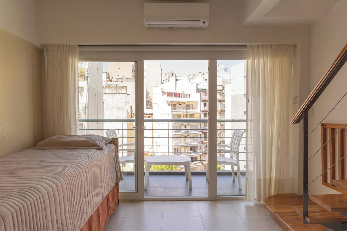 Un dormitorio con cama, un balcón con silla y una escalera.