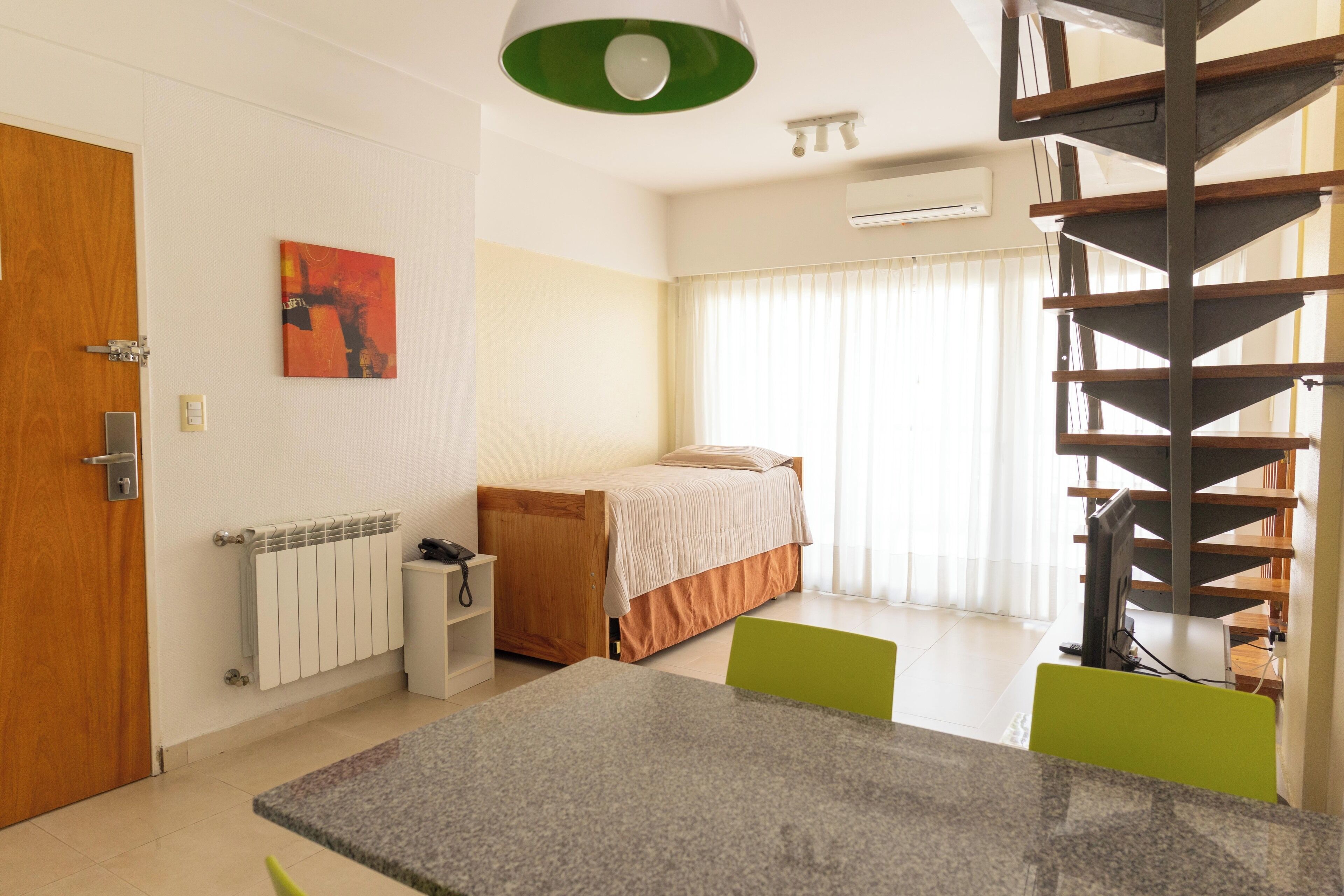 A modern bedroom with a bed, a desk, and a chair. There is a window with curtains, a radiator, and a green light fixture.