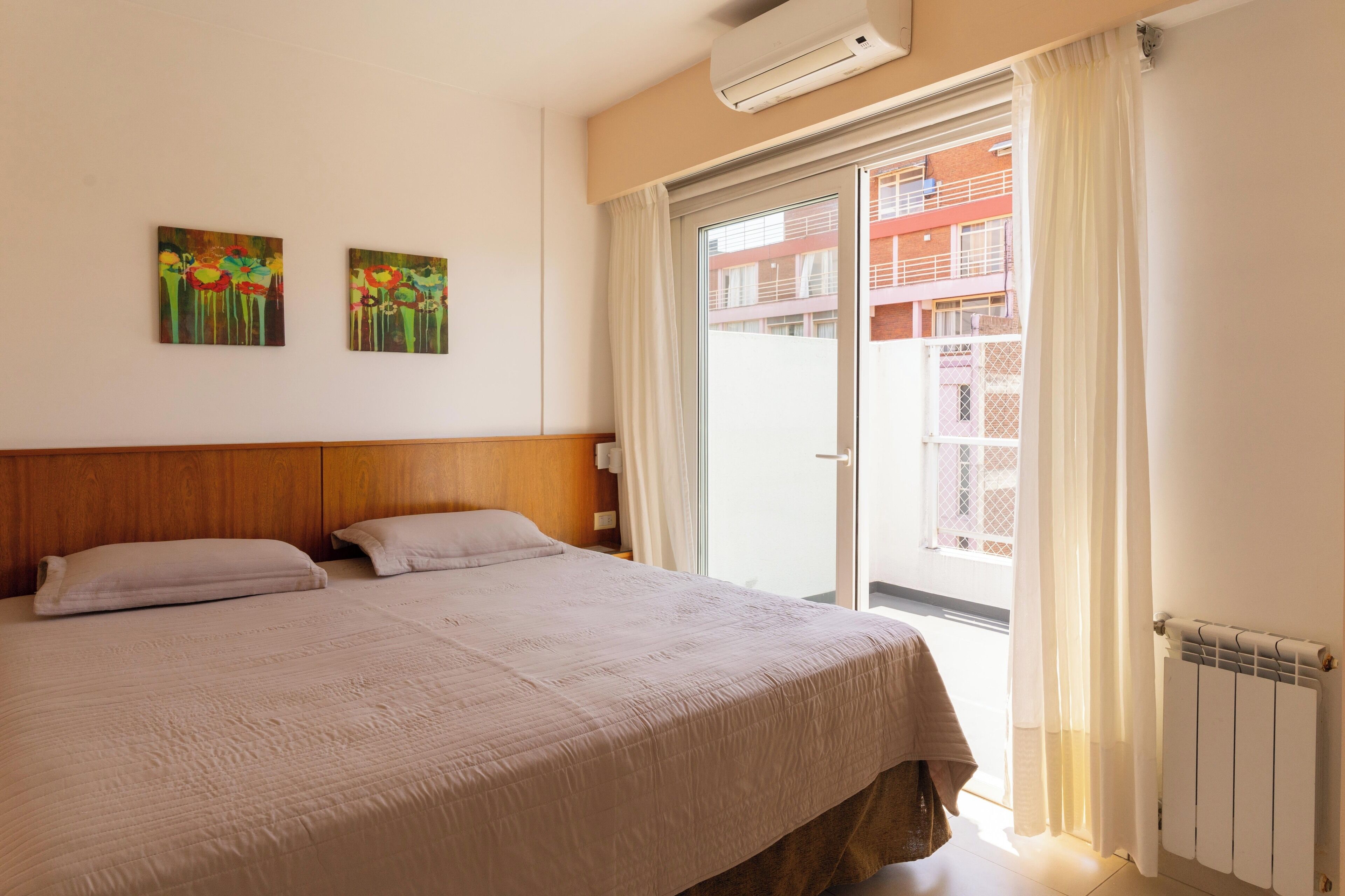 A bedroom with a bed, two wall-mounted paintings, a window with curtains, and a radiator.